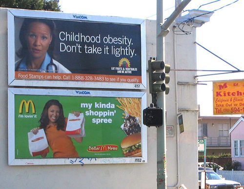 Advertisement placards on the street warning about the dangers of childhood obesity, and below an ad for McDonalds